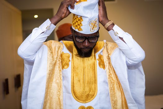 A man adjusts his traditional Nigerian agbada, featuring intricate embroidery indoors.